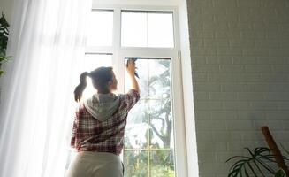 Frau manuell wäscht das Fenster von das Haus mit ein Lappen mit sprühen Reiniger und Mopp Innerhalb das Innere mit Weiß Vorhänge. Wiederherstellen bestellen und Sauberkeit im das Frühling, Reinigung warten foto