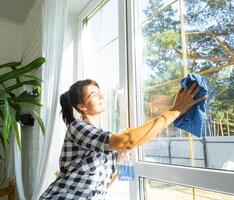 Frau manuell wäscht das Fenster von das Haus mit ein Lappen mit sprühen Reiniger und Mopp Innerhalb das Innere mit Weiß Vorhänge. Wiederherstellen bestellen und Sauberkeit im das Frühling, Reinigung warten foto