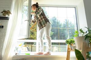 Frau manuell wäscht das Fenster von das Haus mit ein Lappen mit sprühen Reiniger und Mopp Innerhalb das Innere mit Weiß Vorhänge. Wiederherstellen bestellen und Sauberkeit im das Frühling, Reinigung warten foto