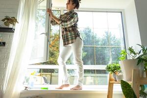 Frau manuell wäscht das Fenster von das Haus mit ein Lappen mit sprühen Reiniger und Mopp Innerhalb das Innere mit Weiß Vorhänge. Wiederherstellen bestellen und Sauberkeit im das Frühling, Reinigung warten foto