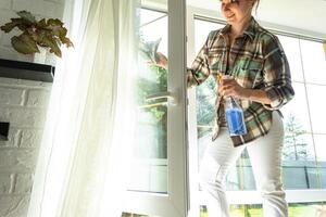 Frau manuell wäscht das Fenster von das Haus mit ein Lappen mit sprühen Reiniger und Mopp Innerhalb das Innere mit Weiß Vorhänge. Wiederherstellen bestellen und Sauberkeit im das Frühling, Reinigung warten foto