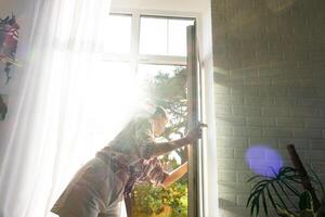 Frau manuell wäscht das Fenster von das Haus mit ein Lappen mit sprühen Reiniger und Mopp Innerhalb das Innere mit Weiß Vorhänge. Wiederherstellen bestellen und Sauberkeit im das Frühling, Reinigung warten foto