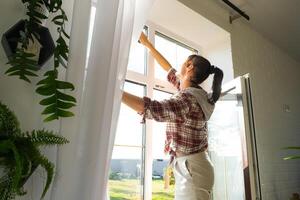 Frau manuell wäscht das Fenster von das Haus mit ein Lappen mit sprühen Reiniger und Mopp Innerhalb das Innere mit Weiß Vorhänge. Wiederherstellen bestellen und Sauberkeit im das Frühling, Reinigung warten foto