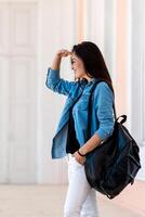 ziemlich Frau mit Rucksack. Aussicht von jung Frau gekleidet im Blau Hemd und Weiß Jeans. suchen beim Seite mit Hand in der Nähe von Augen. Aussicht von das Seite. halbes Gesicht Sicht. foto