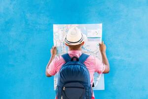 Mann mit ein traurig Reise Hut und Rucksack, suchen beim ein Karte, im Vorderseite von ein Blau Straße Mauer. foto