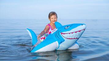 glücklich Mädchen von europäisch Aussehen Alter von 7 Schwimmen auf ein aufblasbar groß Hai Spielzeug im das Meer.Familie Sommer- Berufung Konzept. foto
