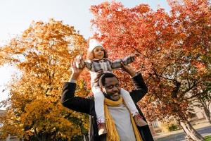 schwarzes Mädchen, das Spaß hat und im Herbstpark am Hals seines Großvaters sitzt foto