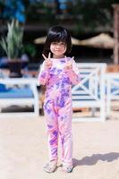 vertikal abgebildet. glückliches asiatisches süßes Mädchen, das steht und postet, machen Sie ein Foto. Kind, das in die Kamera schaut. süßes Lächeln. am Strand. Sommerzeit. Kinder, die Badeanzug tragen. Baby im Alter von 4-5 Jahren.