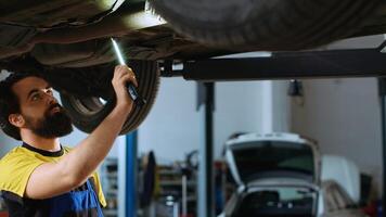 präzise Mechaniker unterhalb Fahrzeug auf Overhead Aufzug im Garage, Überprüfung Komponenten. Mitarbeiter mit Arbeit Licht zu machen sicher Automobil Unterboden ist im perfekt Zustand, tun schließen Untersuchung foto