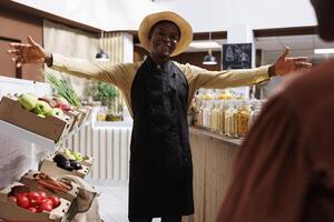 ein modern Öko freundlich Supermarkt im Besitz durch ein freundlich afrikanisch amerikanisch Mann tragen ein Hut und ein Schürze. er verkauft ein Vielfalt von frisch und gesund Produkte, einladend Kunden zu Geschäft mit öffnen Waffen. foto