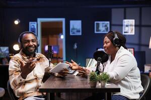 bärtig Mann lacht während ein Komödie Podcast wie das Gastgeber Witze um mit afrikanisch amerikanisch weiblich Blogger. männlich Journalist unterhält beide das Gast und Publikum Aufpassen von heim. foto