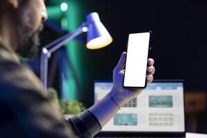Mann vertikal halten Handy, Mobiltelefon Gerät mit isoliert Copyspace Attrappe, Lehrmodell, Simulation Vorlage. im Vorderseite von ein Laptop Bildschirm, ein männlich Individuell ist greifen ein Smartphone zeigen leer Weiß Bildschirm. foto