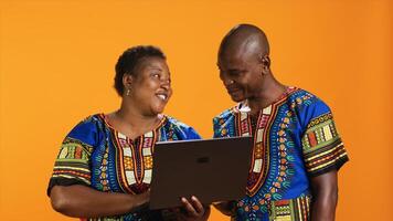 lächelnd afrikanisch amerikanisch Menschen aussehen beim Webseite auf Laptop, Scrollen durch Sozial Medien Netzwerk und Surfen Netz. verheiratet Paar im traditionell Kleidung mit pc zu prüfen online Webseiten, Studio. foto
