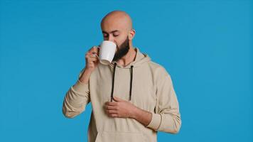 Muslim Person genießen frisch Koffein Aroma im Studio, Trinken Tasse von Kaffee und Stehen Über Blau Hintergrund. Mitte östlichen Erwachsene riechen Getränk und nehmen ein Schluck. Kamera 1. foto