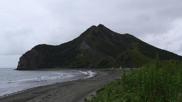 Aussicht von schön Strand mit Grün Gras und Berg. Clip. malerisch Landschaft mit Grün Gras auf Hintergrund von Meer Küste mit Berg. Küste von Meer mit Sand und felsig Berg auf wolkig Tag foto