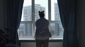 Rückseite Aussicht von ein Frau mit gefaltet Hände suchen aus das Fenster im inländisch Zimmer. Medien. jung nachdenklich weiblich im Zuhause beiläufig Kleider Stehen in der Nähe von groß Fenster beim heim. foto