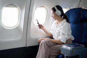 asiatisch jung Frau tragen Kopfhörer Hör mal zu zu Musik- beim zuerst Klasse auf Flugzeug während Flug, Reisen und Geschäft Konzept foto