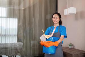 Reinigung Produkt, und Korb mit Frau asiatisch Mitarbeiter Reinigung Bedienung im Schlafzimmer zu sauber Bakterien beim Wohnung. Reiniger oder Maid mit Container zu Arbeit im Zimmer foto