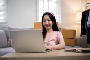 glücklich jung asiatisch Geschäft Inhaber Frau bereiten Paket Box und prüfen online bestellen von Produkt zum liefern zu Kunde auf Laptop Computer. Einkaufen online Konzept foto