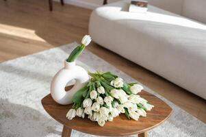 Weiß Tulpen im modern Vase auf hölzern Tabelle mit Sofa im das Hintergrund. foto