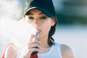 ziemlich jung Hipster asiatisch Frau im schwarz Hut Dampfen E-Zigarette, vaping Gerät beim das Sonnenuntergang. getönt Bild. Nahaufnahme. foto