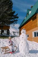 wenig lächelnd Mädchen sitzt auf ein Schlitten in der Nähe von ein Schneemann in der Nähe von ein hölzern Chalet foto