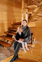 Mutter mit ein wenig Mädchen auf ihr Runde sitzt auf ein Stuhl im ein hölzern Chalet in der Nähe von das Treppe foto