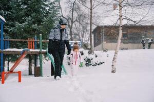 Mama und wenig Tochter gehen halten Hände Vergangenheit ein schneebedeckt rutschen unter Schneefall foto