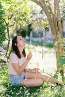 jung Mädchen sitzt unter ein Baum auf das Rasen und weht Seife Luftblasen foto