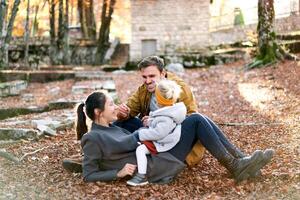 lächelnd Papa erstreckt sich ein Zweig während Sitzung Nächster zu Mama mit ein wenig Mädchen auf ihr Bauch auf gefallen Blätter im das Wald foto