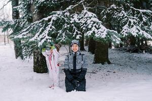 lächelnd Mutter im ein Ski passen sitzt auf ihr Knie im das Schnee in der Nähe von ein klein Kind Stehen in der Nähe von ein schneebedeckt Kiefer Baum foto