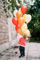 wenig Mädchen Umarmungen ein Bündel von Luftballons im das Garten und sieht aus hoch. zurück Aussicht foto