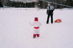 wenig Mädchen geht zu ihr Mutter Stehen mit ein Schlitten im das Schnee. zurück Aussicht foto