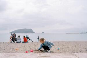 wenig Mädchen gräbt ein Loch im das Sand während hocken gegen das Hintergrund von Mütter mit Kinder Sitzung auf das Ufer foto