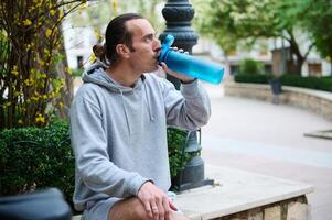 kaukasisch jung Mann Trinken Wasser von ein Sport Flasche im das Stadt foto