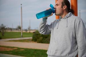 ein Sportler Trinken Wasser von das Flasche, feuchtigkeitsspendend und ruhen nach schwer trainieren auf das städtisch Sportplatz foto