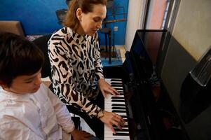 gut aussehend Jugendlicher Junge durchführen klassisch Melodie auf großartig Klavier. kaukasisch Teenager Pianist spielen Klavier unter das Orientierungshilfe von seine Musik- Lehrer beim heim. akustisch Gitarre hängend auf ein Blau Mauer foto