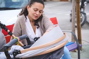 Nahansicht Porträt von schön jung Mutter Sitzung auf städtisch Park Bank mit ihr Neugeborene Baby Schlafen im das Baby Kinderwagen foto