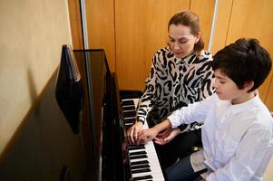 authentisch Porträt von ein Frau Pianist Musiker halten das Hände von ein Teenager Junge, zeigen das wahr Position von Finger auf Klavier Schlüssel, erklären das Klavier Lektion während Individuell Musik- Klasse beim Zuhause foto