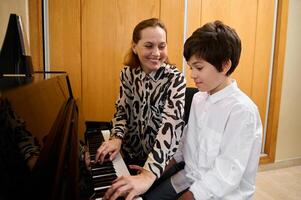 lächelnd liebend Mutter spielen Klavier, Sitzung in der Nähe von seine Jugendlicher Sohn, durchführen Weihnachten Sohn, genießen das Rhythmus von Melodie foto