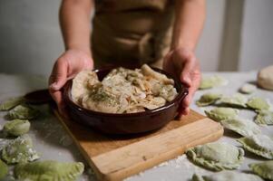 Nahansicht von das Hände von Hausfrau Portion köstlich hausgemacht Knödel im ein Lehm Gericht, Stehen beim bemehlt Marmor Küche Tabelle mit roh ungekocht Warennyki. hausgemacht Essen Konzept. ethnisch kulinarisch foto