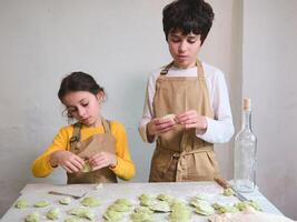 Teenager Junge und Schule Mädchen Kochen zusammen im das Zuhause Küche, Herstellung Knödel, Portion ihr Mama zu bereiten ein Abendessen, Stehen beim bemehlt Tisch, gekleidet im Beige Koch Schürze foto