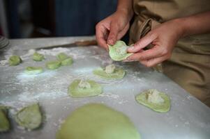 Nahansicht Frau Füllung Teig mit püriert Kartoffeln, Modellieren Knödel, Stehen beim Küche Tabelle mit geknetet Teig und Zutaten. das Prozess von Kochen Ravioli, Wareniki, Vegetarier pelmeni foto