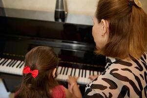 Lernen zu abspielen das Klavier. Rückseite Aussicht von ein weiblich Musiker Pianist geben ein Klavier Lektion zu ein wenig Schülerin während Individuell Musik- Klasse foto