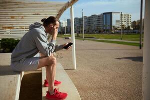 sportlich Mann tragen Kopfhörer, Überprüfung Handy, Mobiltelefon App auf seine Smartphone, Surfen Neu Fitness Anwendung, Sitzung auf Stein Bank foto