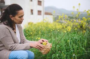 zuversichtlich jung Frau Versammlung Kräuter und Pflanzen zum medizinisch verwenden, Sammeln Blumen im ein Papier Tasche im das Natur foto