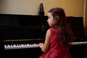 zuversichtlich kaukasisch elegant Schule Mädchen im rot Kleid, Sitzung auf ein Stuhl und durchführen klassisch Musical Komposition, berühren das Schlüssel und Gefühl Rhythmus während spielen großartig Klavier beim Zuhause foto