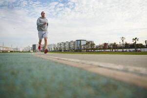 gut aussehend Sportler, sportlich Mann Laufen auf das Brücke auf sonnig Tag, genießen seine Morgen Joggen auf das Promenade foto