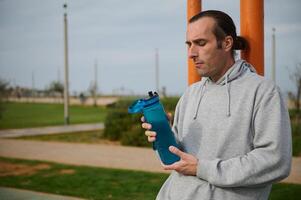 sportlich Mann halten ein Flasche von isotonisch Wasser, ruhen nach schwer trainieren auf das städtisch Sportplatz. Menschen und Sport foto
