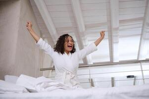 glücklich aufgeregt jung Frau Dehnen im Bett während aufwachen oben im das Morgen, genießen ihr Wochenende beim Zuhause foto